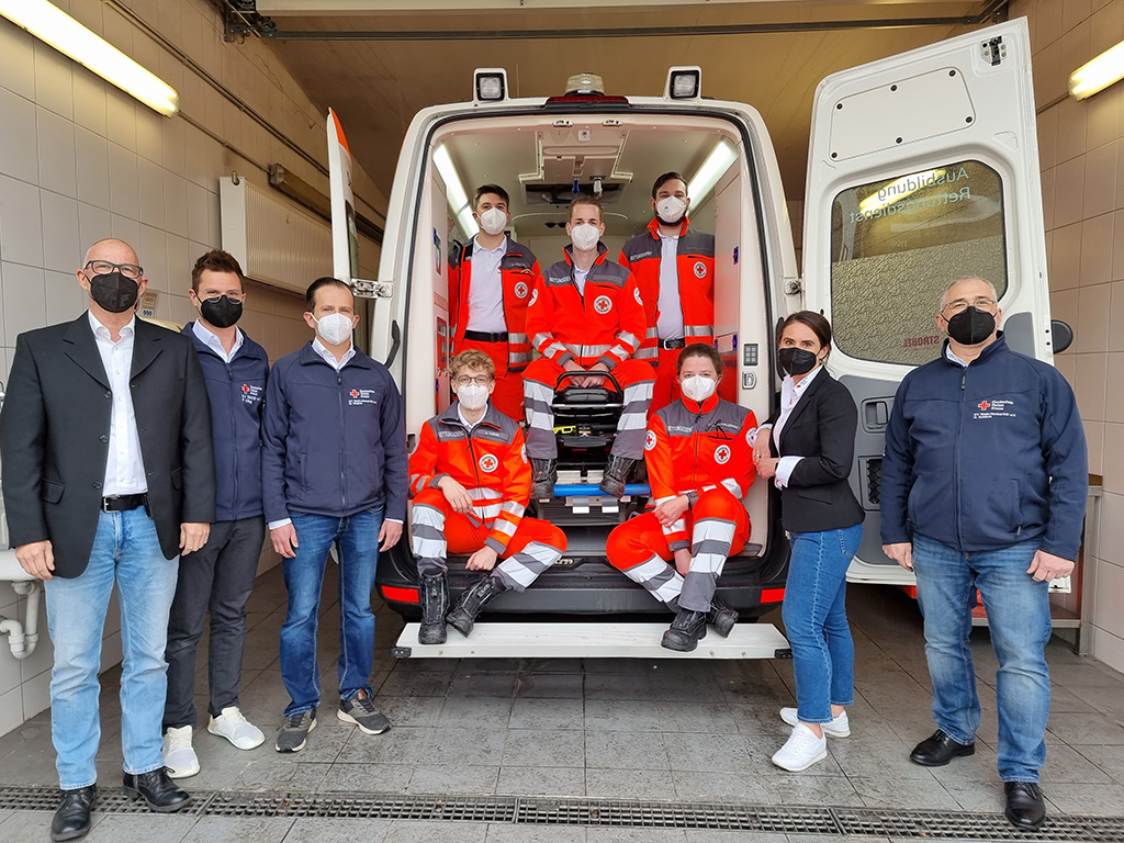 Die fünf neuen Azubis - sitzend im geöffneten Rettungswagen - mit dem Team Ausbildung Rettungsdienst 