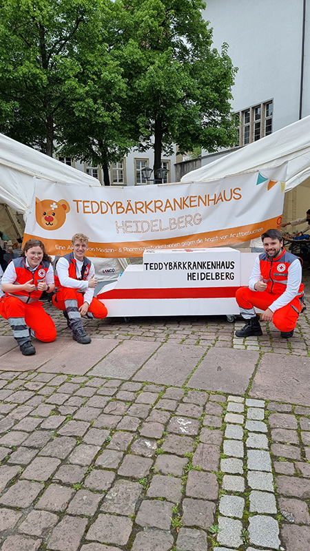 Drei DRK Notfallsanitäter/innen vor einem Banner mit der Aufschrift Teddybärenkrankenhaus Heidelberg