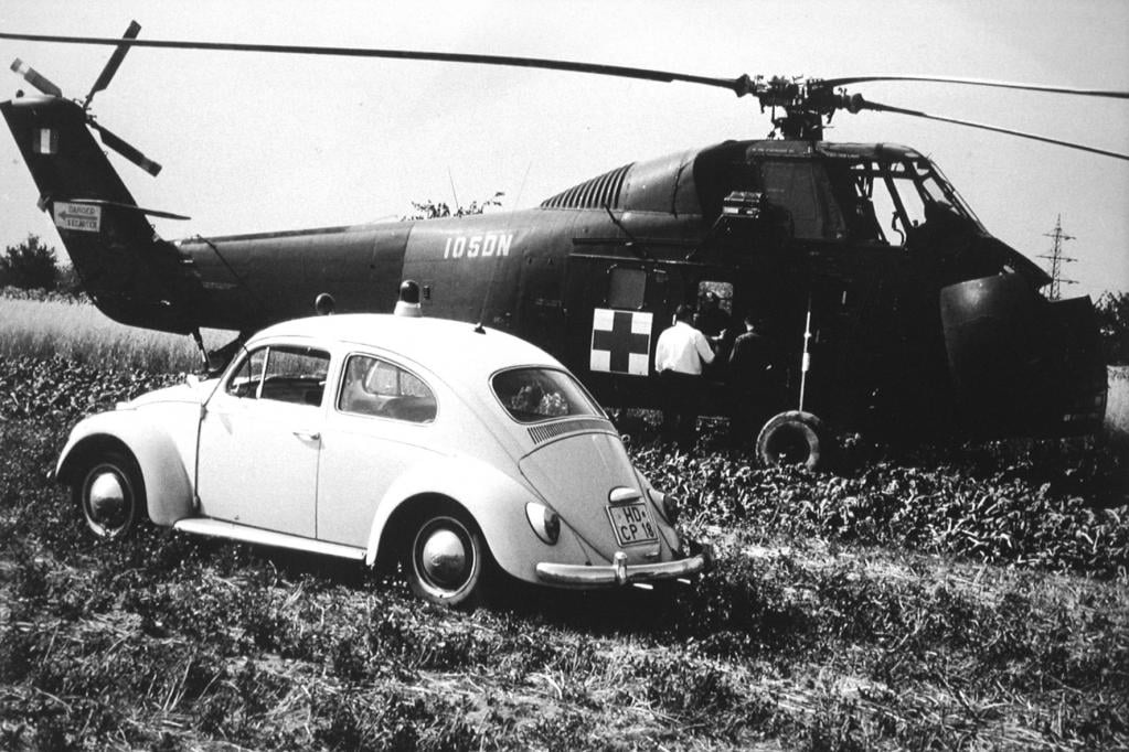 Ein mit Blaulicht ausgestatteter VW Käfer mit dem Namen HD-10 vor einem Rettungshubschrauber