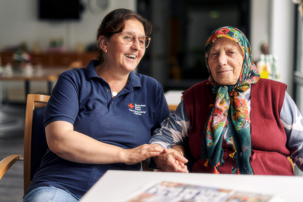 DRK-Mitarbeiterin hält einer Seniorin mit Kopftuch die Hand und unterhält sich mit ihr