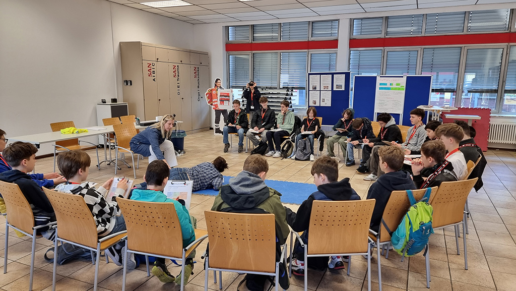Viele Jungen und Mädchen sitzen in einem Kreis auf Stühlen und hören den Erklärungen bezüglich Erste Hilfe zu.