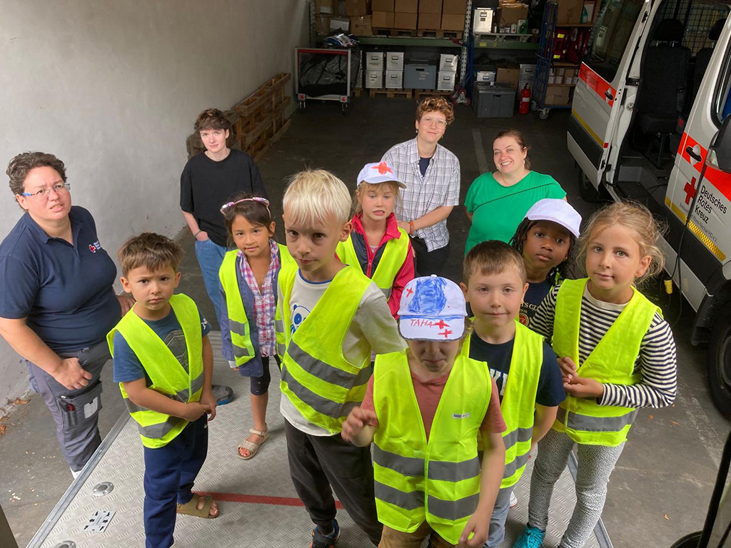 Bild größer ansehen Gruppenbild mit Betreuerinnen und 8 Kindern neben einem Rettungswagen
