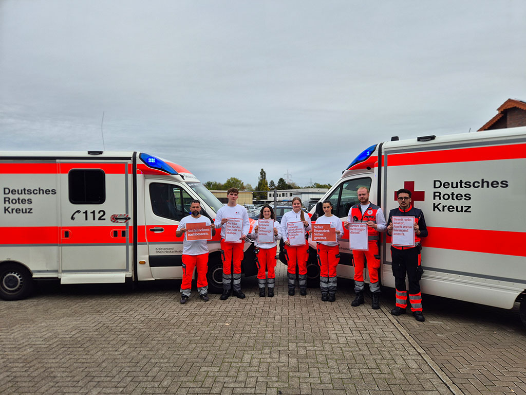 Team der Rettungswache vor zwei Rettungswagen halten Zettel hoch, auf denen sie aufmerksam machen, dass die Notfallreform nachgebessert werden soll.