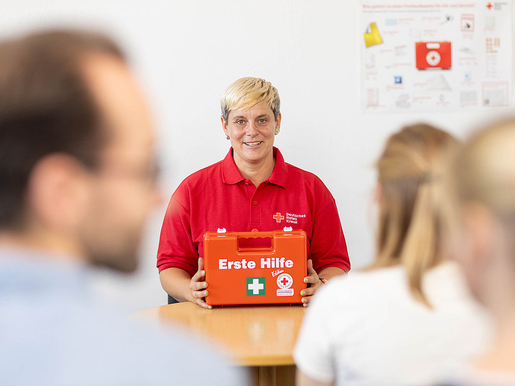 Junge Frau mit rotem DRK-Shirt zeigt Kursteilnehmern einen Erste Hilfe Koffer