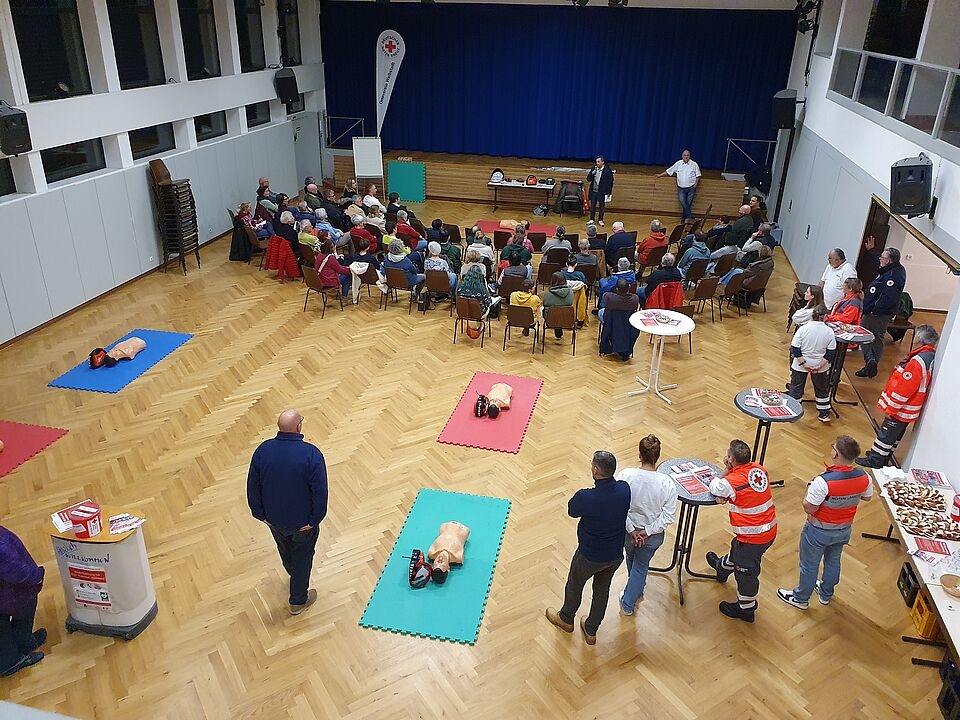 Blick in die Halle: viele Teilnehmer sitzen auf Stühlen und hören den Vortrag des Bürgermeisters, hinten sind Matten mit Puppen für die Übung der Wiederbelebung vorbereitet