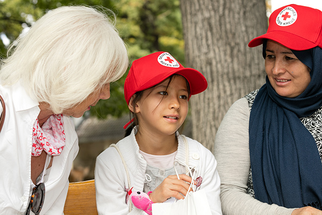 DRK-Mitarbeiterin unterhält sich mit einer ausländischen Mutter und ihrer Tochter. Beide tragen rote Schildmützen mit DRK-Logo.
