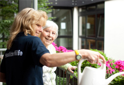 Frau gießt bei einer Seniorin Blumen auf dem Balkon