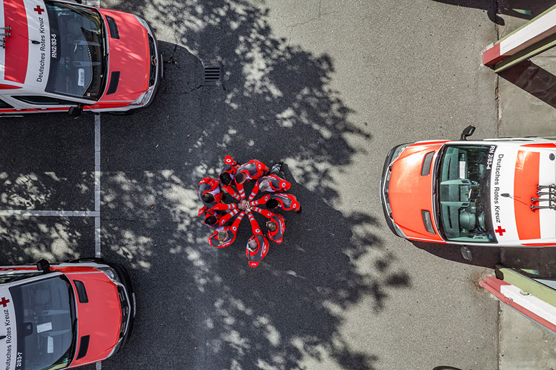 Drei Fahrzeuge auf einem Hof von oben fotografiert. In der Mittte stehen DRK-Mitarbeiter in einem Kreis und halten ihre Hände in der Mitte aufeinander